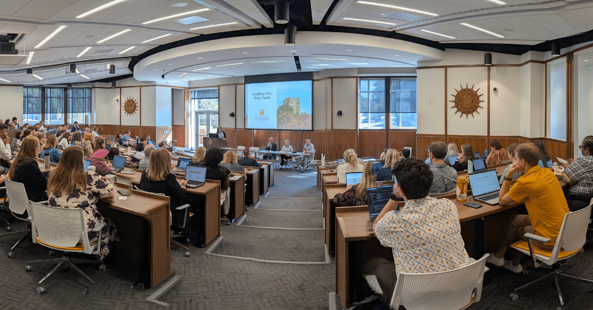 This image depicts a tiered, lecture-style room filled with people sat in seats and facing the front of the room. At the front of the room are a lectern and table, with one person behind the lectern and three people sat behind the table, and a projector screen extended with a presentation slide visible that says "Leading with Data Panel"